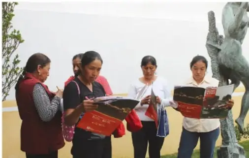 Grupo de personas, en su mayoría mujeres, revisa folletos informativos al aire libre; al fondo se observa una escultura.