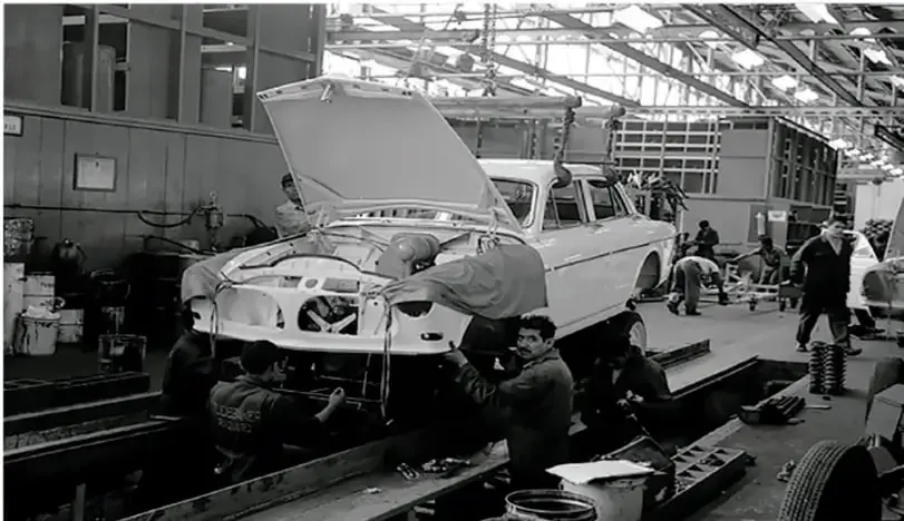 Fotografía en blanco y negro de una línea de ensamblaje automotriz. Varios trabajadores colocan la carrocería de un automóvil sobre el chasis en una fábrica. Se observan herramientas y piezas alrededor.
