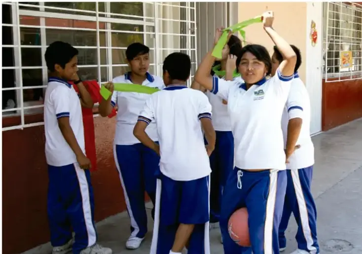 Grupo de estudiantes de secundaria en el patio escolar realizando actividades con cintas de colores (una alumna se coloca una cinta en la cabeza), uno de ellos con un balón de baloncesto; visten uniforme blanco con pantalón azul.