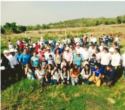 Fotografía de un grupo numeroso de personas de una comunidad reunidas al aire libre en un campo verde, con adultos y niños.