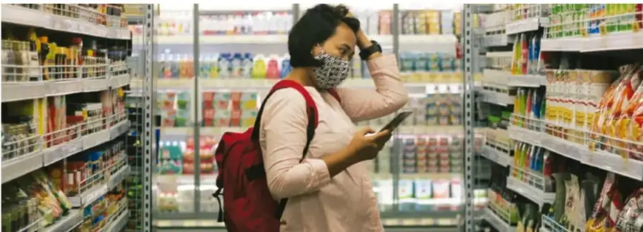 Mujer con cubrebocas y mochila roja observando productos en los pasillos de un supermercado.