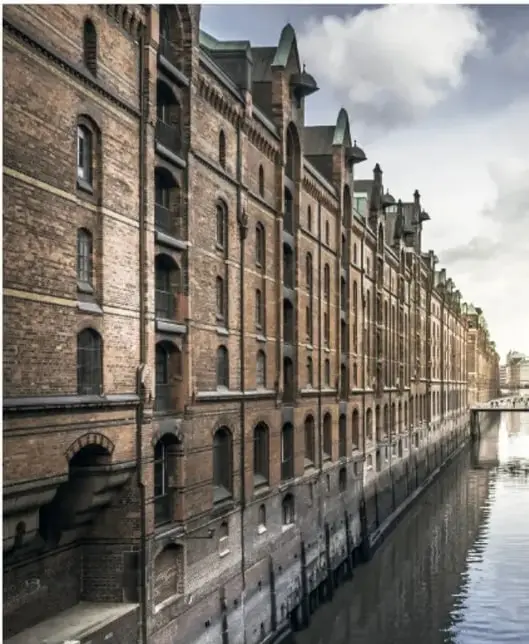 Fotografía de un edificio de ladrillo de varias plantas, con hileras de ventanas arqueadas, situado junto a un canal donde se refleja.