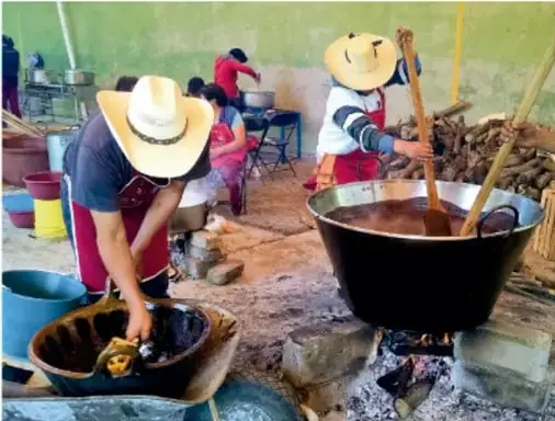 Personas con sombreros de paja preparando comida en ollas grandes sobre fogatas al aire libre, en una actividad comunitaria de cocina tradicional.