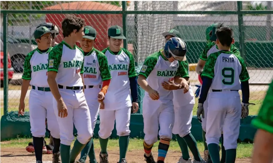 Fotografía de un equipo juvenil de béisbol con camisetas blancas y verdes (que dicen BAAXAL) y gorras, reunido en el campo de juego tras una jugada, mostrando camaradería.
