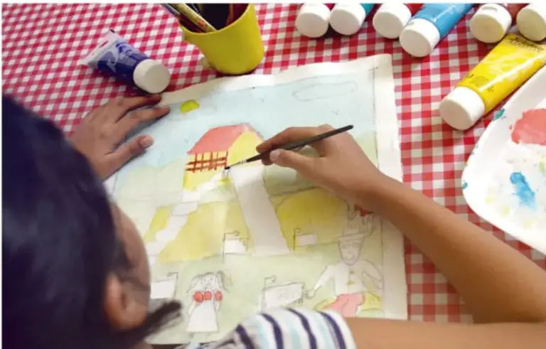 Niña pintando con acuarelas un dibujo de casas y paisaje sobre un mantel de cuadros rojos y blancos, rodeada de tubos de pintura.