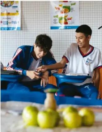 Dos estudiantes de telesecundaria trabajando sentados con carpetas y materiales escolares; al frente hay frutas (manos) sobre la mesa.