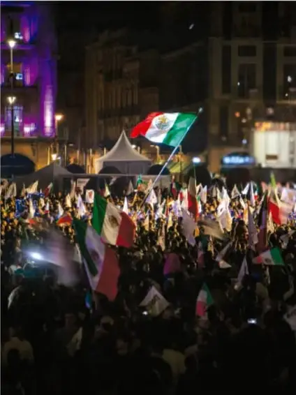 Fotografía de una multitud en una plaza pública, con banderas de México, en un ambiente de manifestación o celebración cívica.