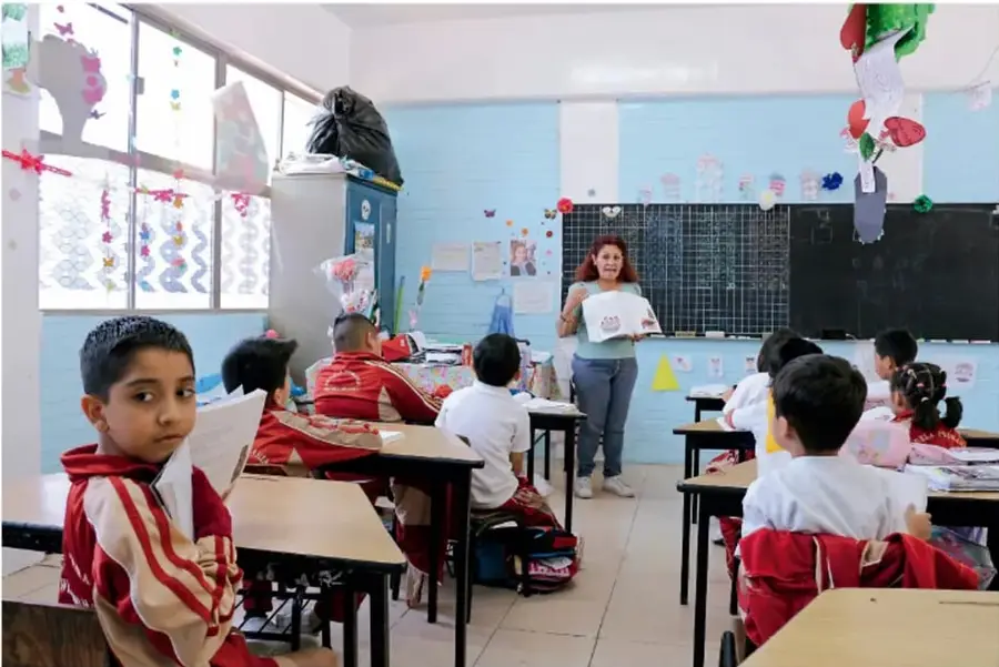 Fotografía de un aula: una maestra frente al grupo sostiene un libro mientras las niñas y los niños atienden sentados en sus pupitres; pizarrón, trabajo artesanal y material escolar decoran el salón.