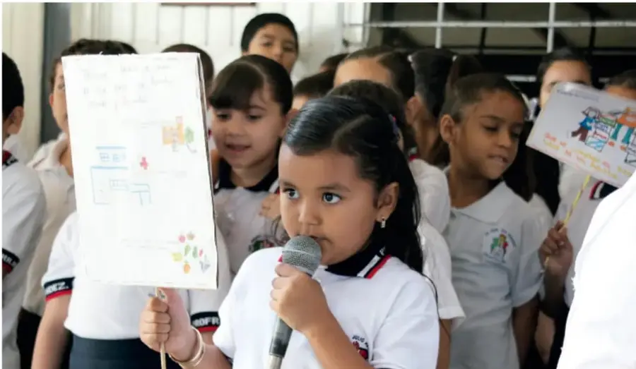 Fotografía de una estudiante de primaria hablando por micrófono frente a sus compañeros, sosteniendo un cartel con un dibujo; los demás alumnos llevan uniforme blanco y escuchan.