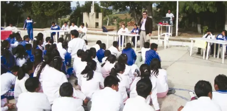 Fotografía de una asamblea escolar al aire libre: estudiantes con uniforme blanco y azul sentados en el piso, escuchando a un orador de pie frente a ellos, con árboles y edificios al fondo.