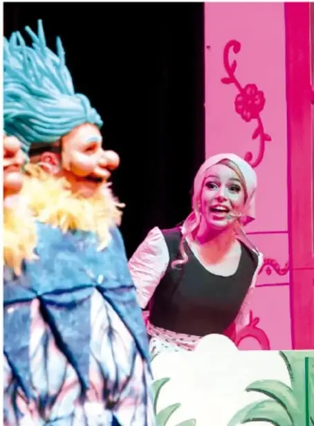 Fotografía de una representación teatral: actores disfrazados, uno con peluca azul y barba, ante una escenografía rosa con motivos decorativos, en escena de un cuento.