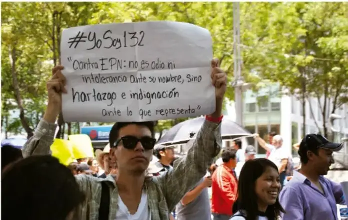 Fotografía de una manifestación callejera del movimiento #YoSoy132 en México; un joven con lentes de sol sostiene una pancarta escrita a mano que dice: '#YoSoy132. Contra EPN: no es odio ni intolerancia ante su nombre, sino hartazgo e indignación ante lo que representa'. Alrededor hay otros manifestantes.