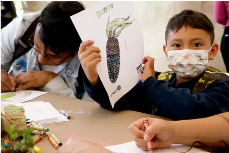Fotografía de estudiantes en un salón: una niña dibuja y un niño con cubrebocas muestra un cartel con el dibujo de un elote y la palabra 'maíz'.
