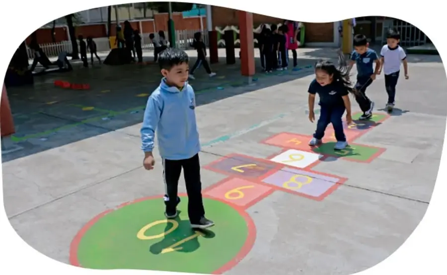 Fotografía de niñas y niños jugando avión (rayuela) en el patio escolar, sobre casillas numeradas del 10 al 4 pintadas de colores en el piso; un niño camina junto al inicio del tablero.