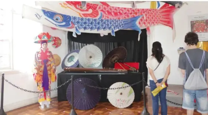 Fotografía de una exposición artística multicultural: koinobori (carpes japoneses) colgados, vestido tradicional japonés, sombrillas de papel japonesas sobre un pedestal y dos estudiantes observando la muestra.