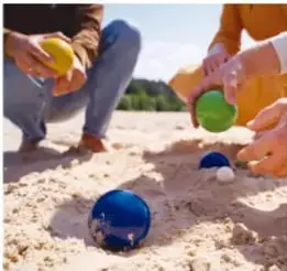 Manos de niños jugando con canicas sobre arena; una canica azul en reposo y otra verde sostenida en la mano.