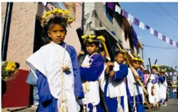 Fotografía de niñas y niños con vestimenta tradicional y coronas de flores participando en un desfile o celebración comunitaria en la calle.