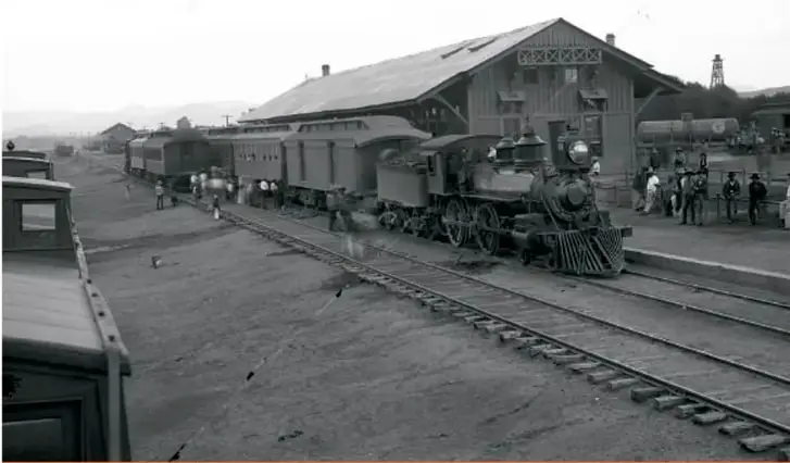 Fotografía antigua de una estación de ferrocarril con una locomotora de vapor y varios vagones; hay personas en el andén.