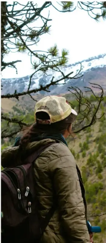 Persona con mochila y gorra observando un paisaje montañoso con árboles, en actividades al aire libre.