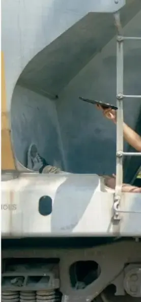 Fotografía de una persona asomada desde la estructura metálica de un tren (escalerilla/vagón), en tonos grises; se relaciona con los migrantes que viajan sobre el tren.
