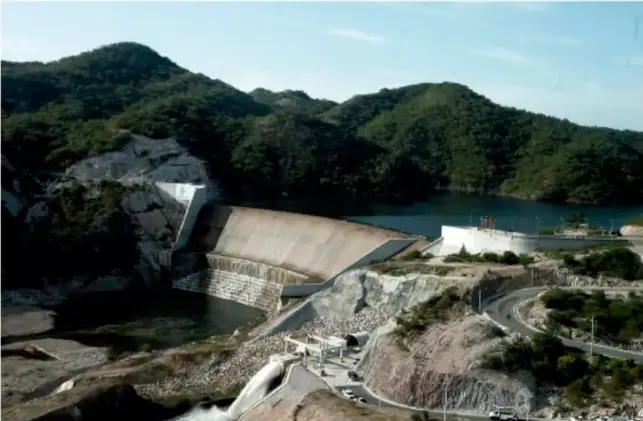 Fotografía aérea de una presa hidroeléctrica en una zona montañosa boscosa, con el embalse y la estructura de concreto visibles.