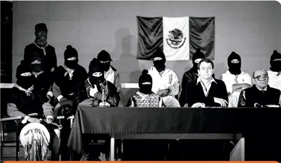 Fotografía en blanco y negro de una mesa de diálogo por la paz entre el gobierno federal y el Ejército Zapatista de Liberación Nacional en la catedral de San Cristóbal de las Casas, Chiapas. Se observan varias personas, algunas con pasamontañas, sentadas tras una mesa, con una bandera de México al fondo.