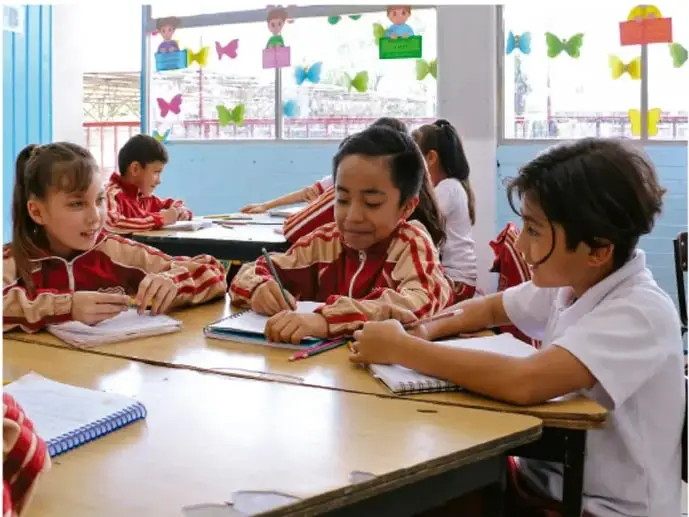 Fotografía de tres niñas y niños trabajando juntos en un pupitre: escriben y conversan; el salón tiene decoraciones de mariposas de papel en la ventana.
