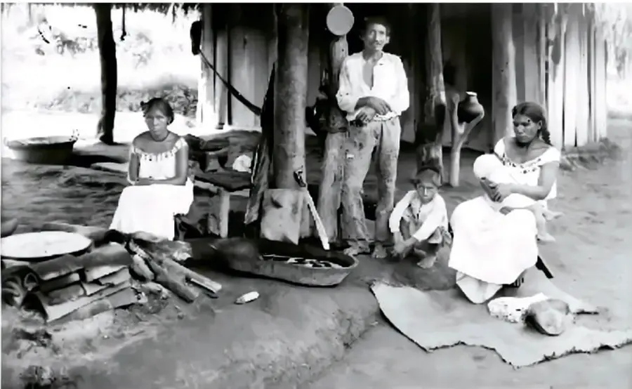 Fotografía en blanco y negro de una familia indígena en un entorno rural, con una mujer sentada en el suelo, un hombre de pie y niños.