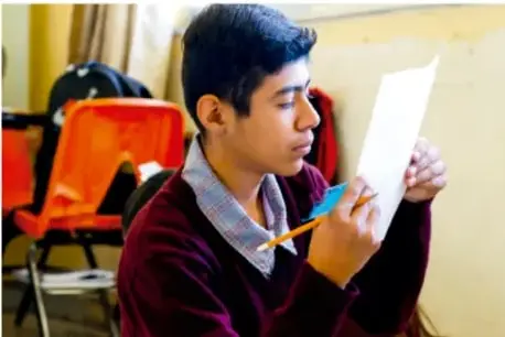 Fotografía de un estudiante de telesecundaria sentado, sosteniendo una hoja de papel y un lápiz, aparentemente revisando o leyendo un texto, con sillas naranjas al fondo.
