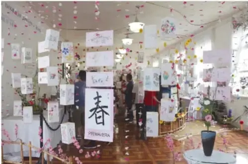 Fotografía de un salón con una exposición artística multicultural: láminas y dibujos colgados con hilo, un cartel que dice 'PRIMAVERA' con un carácter chino, arreglos florales y personas visitando la muestra.