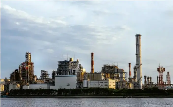 Fotografía de una planta industrial con chimeneas y estructuras metálicas a orillas del agua, al atardecer; representa la generación de energía/electricidad que contamina.