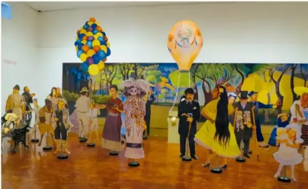 Fotografía de una exhibición escolar con figuras de cartón y papel que representan personas en un salón, con globos y un mural de fondo.