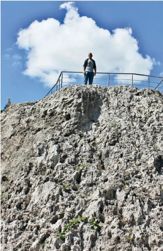 Persona de pie junto al barandal del mirador en la cima del cono rocoso del Cuexcomate, con cielo azul y nubes.