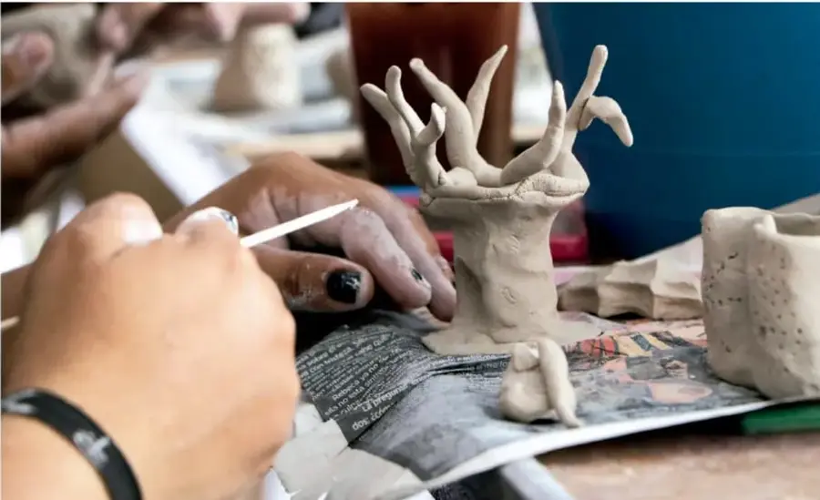 Fotografía de manos trabajando arcilla: una figura de arcilla con forma de árbol o manos entrelazadas sobre un mantel de periódico, en un taller de escultura escolar.