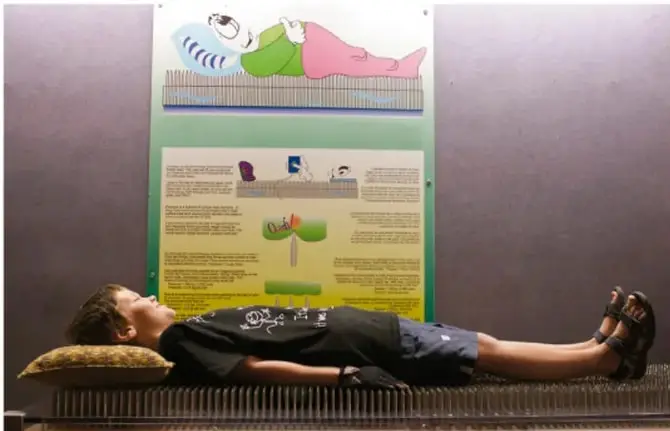 Fotografía de un joven acostado sobre una cama de clavos, con un cartel explicativo al fondo.