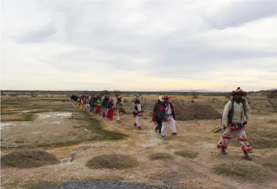 Fotografía de una fila de peregrinos wixaritari con vestimenta tradicional caminando por el desierto de Wirikuta al amanecer o atardecer.