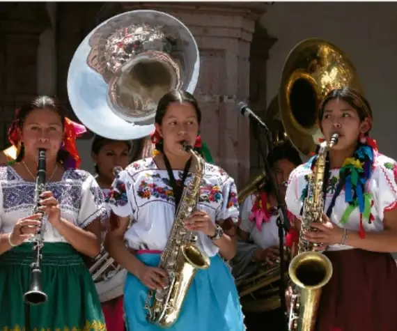 Fotografía de niñas y jóvenes con traje tradicional oaxaqueño tocando clarinete, saxofón y tuba, ilustrando las bandas de viento de las comunidades mixe.