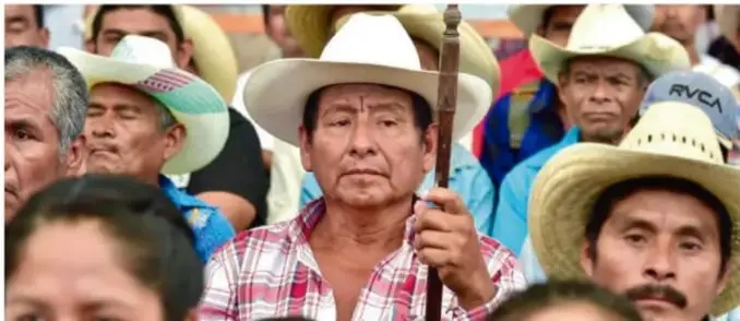 Fotografía de un grupo de personas indígenas adultas, varias con sombreros de ala ancha (sombreros campesinos), una persona al centro porta un bastón; rostros serios, en un acto o reunión comunitaria al aire libre.