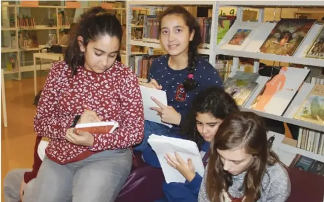 Cuatro estudiantes de telesecundaria sentados en pisos con cojines dentro de una biblioteca, leyendo y tomando notas en cuadernos; hay estantes con libros al fondo.