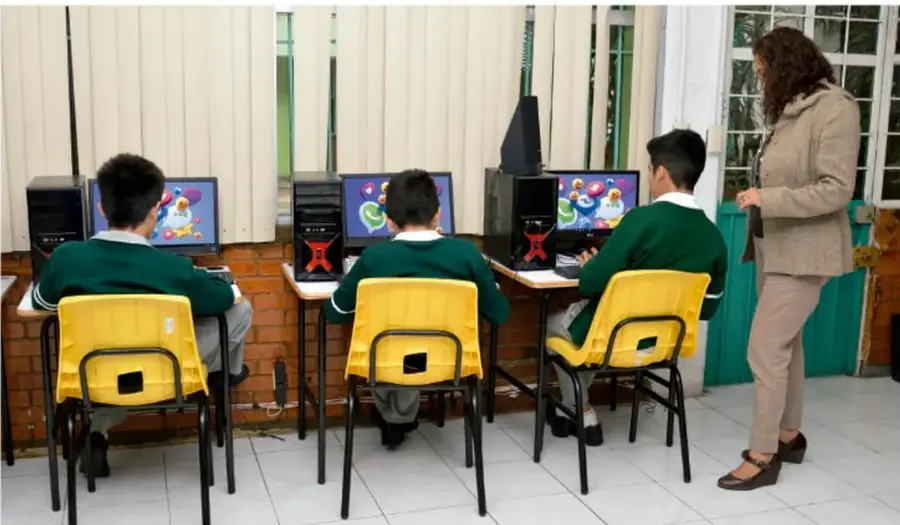 Fotografía de tres estudiantes en green usándo computadoras en el aula mientras una docente los supervisa.