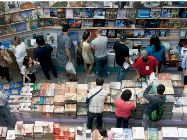 Fotografía de una feria o librería con público joven y adulto observando y seleccionando libros en mesas y estantes, ilustrando la promoción y difusión de la literatura.