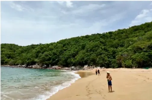 Fotografía de una playa con arena dorada, agua turquesa, colinas boscosas al fondo y algunas personas caminando por la orilla.