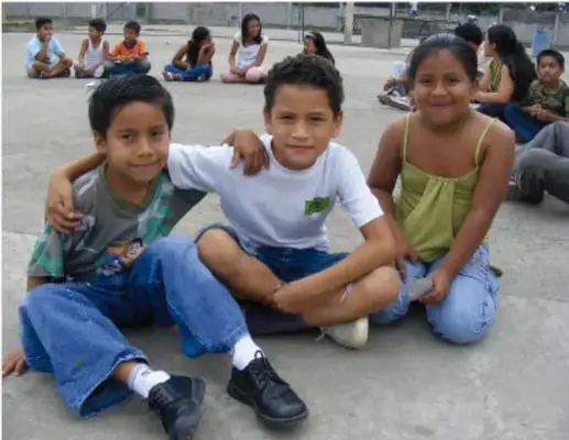 Tres niños sentados en el suelo del patio escolar, abrazados y sonriendo, mientras otros compañeros observan al fondo.