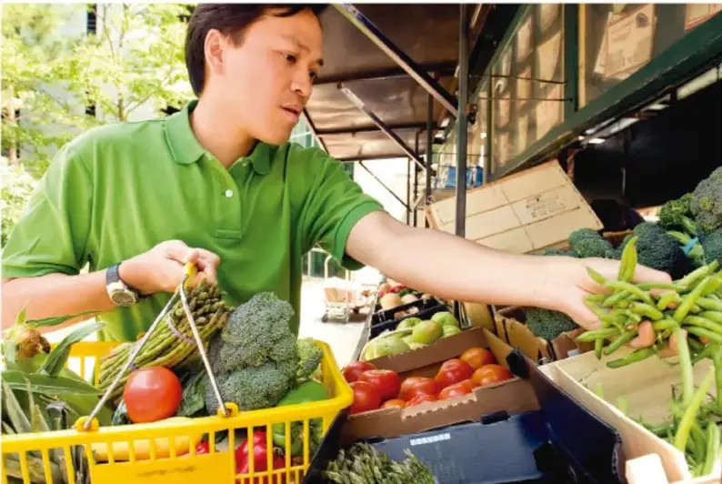 Fotografía de un joven con camisa verde que compra verduras en un mercado: sostiene una canasta amarilla con brócoli, jitomates, elotes y ejotes.