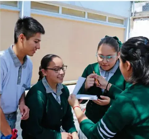 Fotografía de cuatro estudiantes de telesecundaria uniformados (suéter verde) revisando juntos apuntes en un pasillo escolar