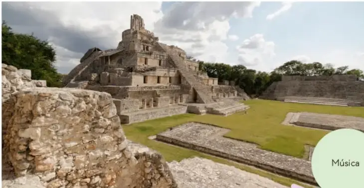 Fotografía de una zona arqueológica con una pirámide y estructuras de piedra de una civilización mesoamericana, rodeada de césped y árboles.