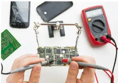Fotografía de una persona manipulando una tarjeta electrónica con un multímetro, acompañada de una pantalla de teléfono rota y una tarjeta verde, ilustrando el uso de metales en dispositivos tecnológicos.