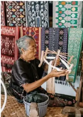 Fotografía de una artesana tejiendo en un telar de cintura frente a textiles con patrones geométricos de colores.