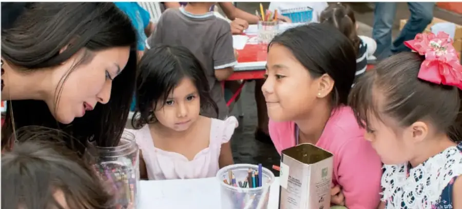 Fotografía de una maestra y varias niñas y niños de primaria trabajando en una mesa con frascos, lápices y una caja, durante una actividad escolar.