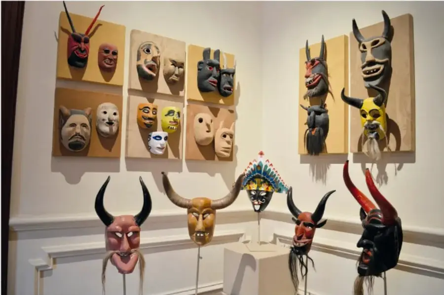 Sala de un museo con muchas máscaras mexicanas expuestas: diablos con cuernos rojos y negros, rostros humanos de madera pintada, máscara amarilla con cuernos de toro y una pieza con penacho multicolor sobre pedestal.